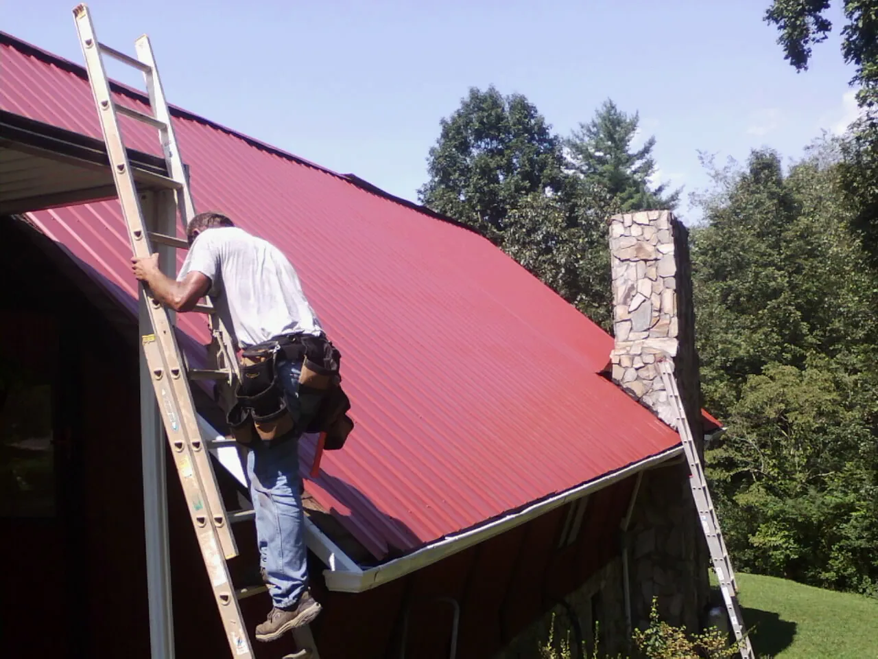 Roofer on a ladder beside a red metal roof and stone chimney — Soffit & Fascia Repair by Roofing Brothers in Chevy Chase
